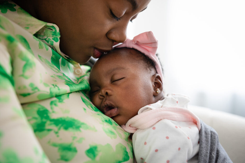 Mother holding her newborn baby in a baby carrier sitting on a sofa bonding with her. They are at their home in Sedgefield, North East England. The baby is sleeping while the mother kisses her head.