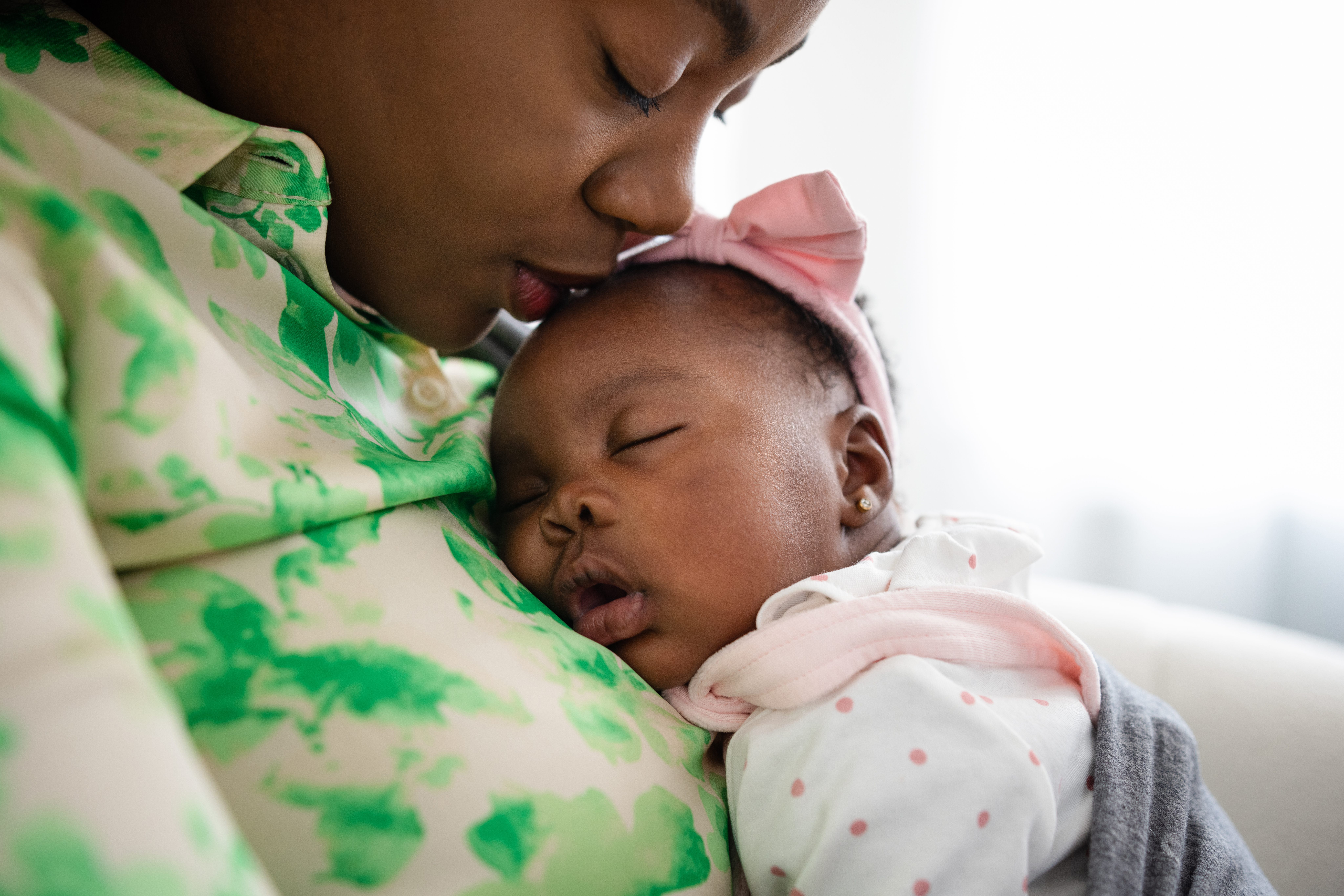 Mother holding her newborn baby in a baby carrier sitting on a sofa bonding with her. They are at their home in Sedgefield, North East England. The baby is sleeping while the mother kisses her head.