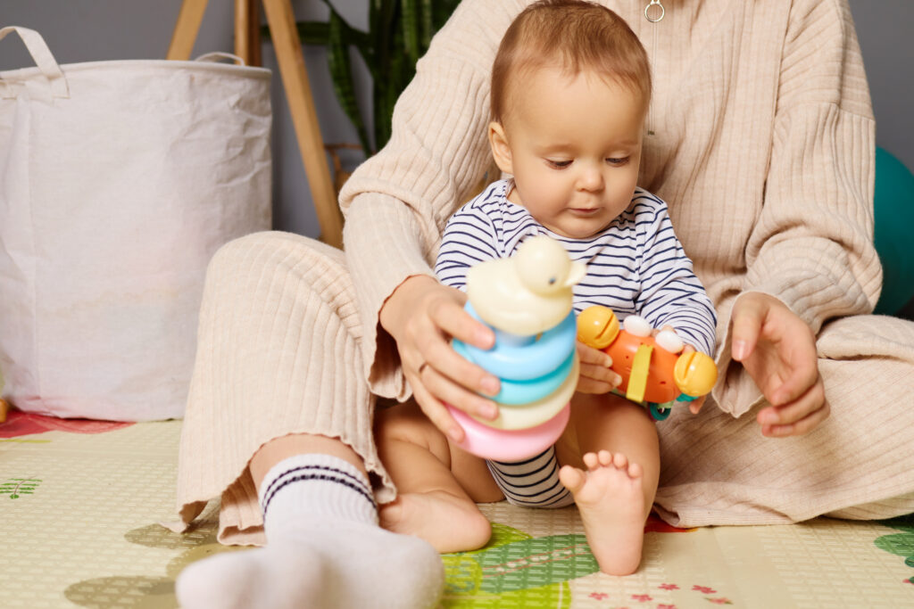 A small child sits on a soft mat, focused on stacking colorful rings and holding a toy, while a caregiver engages nearby in a warm home setting
