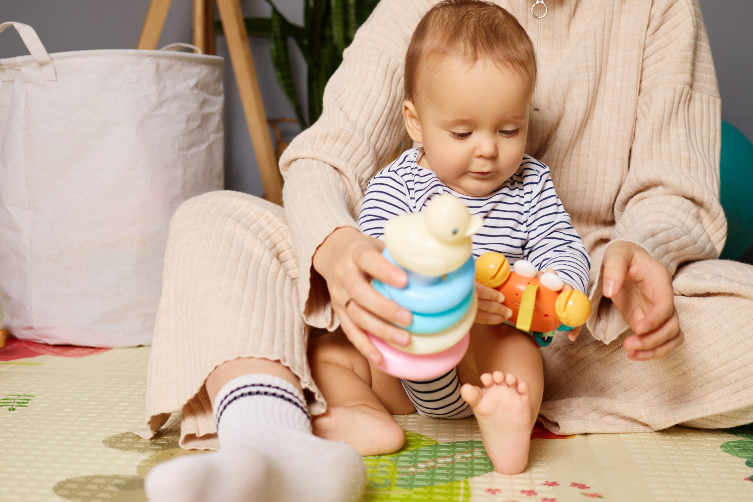 A small child sits on a soft mat, focused on stacking colorful rings and holding a toy, while a caregiver engages nearby in a warm home setting