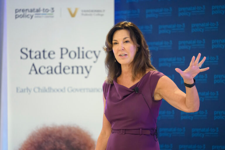 A person in a purple dress speaks at a conference, gesturing with one hand while standing before a backdrop featuring "State Policy Academy" and "prenatal-to-3 policy" branding.