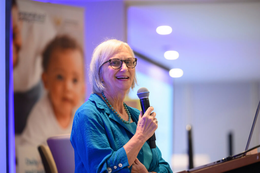 A person with short white hair and glasses, wearing a teal outfit and a beaded necklace, speaks into a microphone while standing at a podium in a well-lit room.