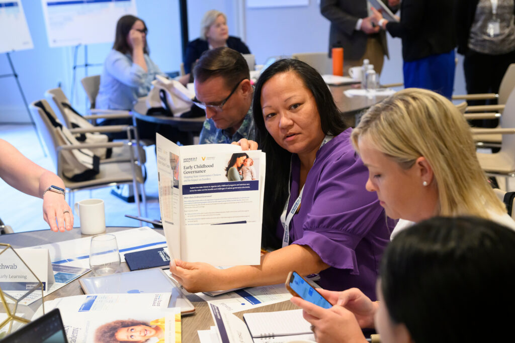 A group of people are gathered around a table, examining a document titled "Early Childhood Governance" during what appears to be a professional meeting or workshop.