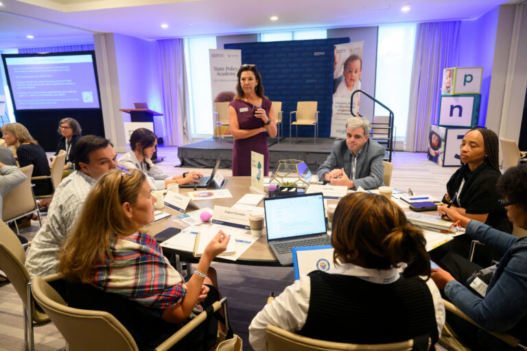 A group of attendees gathers around a table during a State Policy Academy session, attentively listening to a presenter on stage.