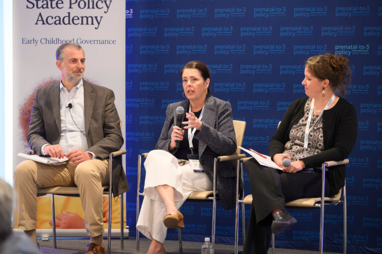 Three individuals participate in a panel discussion at the State Policy Academy, with one speaking into a microphone while the others listen attentively.
