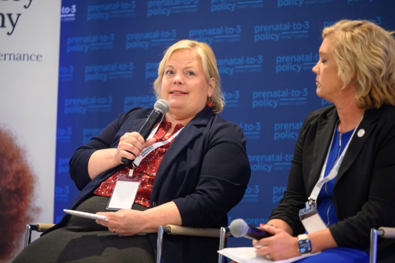 Two individuals engage in a discussion at a conference, with one speaking into a microphone while seated before a branded backdrop.