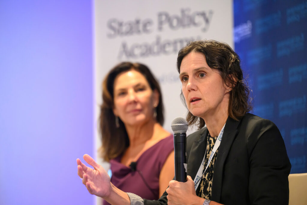 Two individuals are speaking at a State Policy Academy event, with one holding a microphone and gesturing while the other listens attentively.