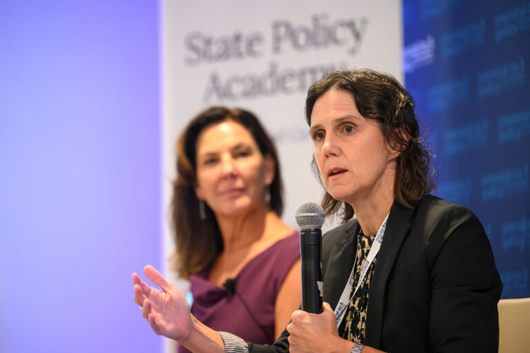 Two individuals are speaking at a State Policy Academy event, with one holding a microphone and gesturing while the other listens attentively.