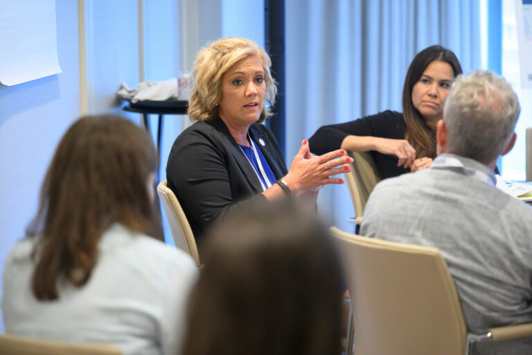 A person with blonde hair speaks animatedly during a meeting, gesturing with hands while others listen attentively around a table.