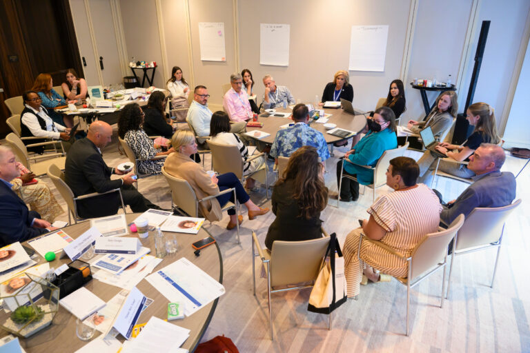 A group of individuals is gathered in a conference room, seated around tables with documents and laptops, engaged in a collaborative discussion.