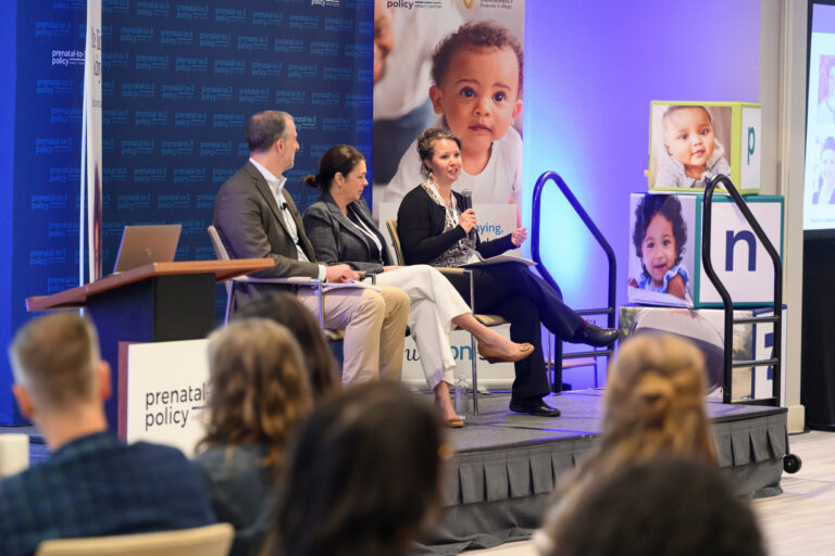 Three individuals sit on a stage at a prenatal-to-3 policy event, with a speaker holding a microphone and a large banner featuring a child's image behind them.