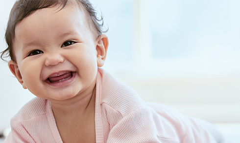 A smiling baby with short, dark hair wearing a light pink outfit, looking directly at the camera. The background is softly blurred, creating a warm, cheerful atmosphere.